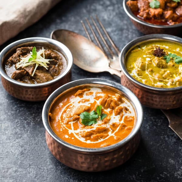 Four copper bowls filled with various Indian curries, garnished with herbs and spices, on a dark surface.