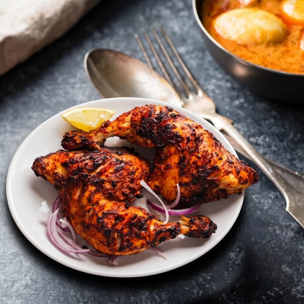 Two grilled chicken legs with lemon and red onions on a white plate, with a curry bowl.